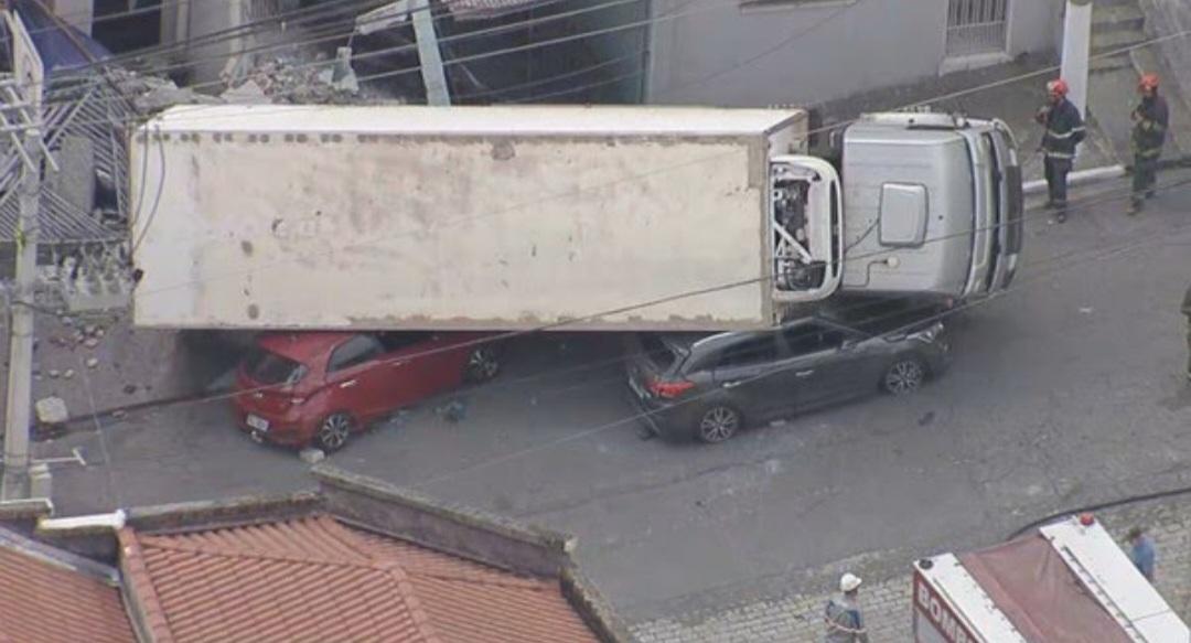 Caminhão baú atinge muro de residência e tomba em cima de dois carros 